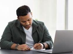 一个人坐在桌边，用笔记本电脑在纸上写东西